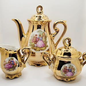 May include: A gold-colored tea set featuring a teapot, creamer, and sugar bowl. Each piece has a decorative handle and a small, oval-shaped image of a couple in period clothing. The set is likely vintage and suitable for serving tea.