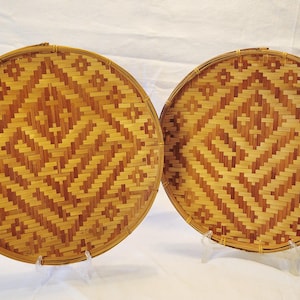 May include: Two round, woven bamboo trays with a geometric diamond pattern in shades of light and dark brown. The trays are displayed on clear stands against a white background. The trays are likely used for serving food or as decorative items.