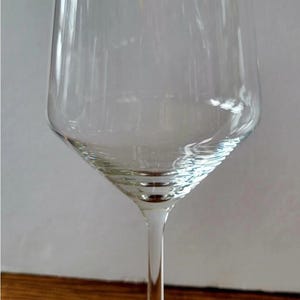 May include: A clear, stemmed wine glass with a wide, slightly tapered bowl and a thin stem. The glass sits on a round base. The glass is empty and reflects the light.