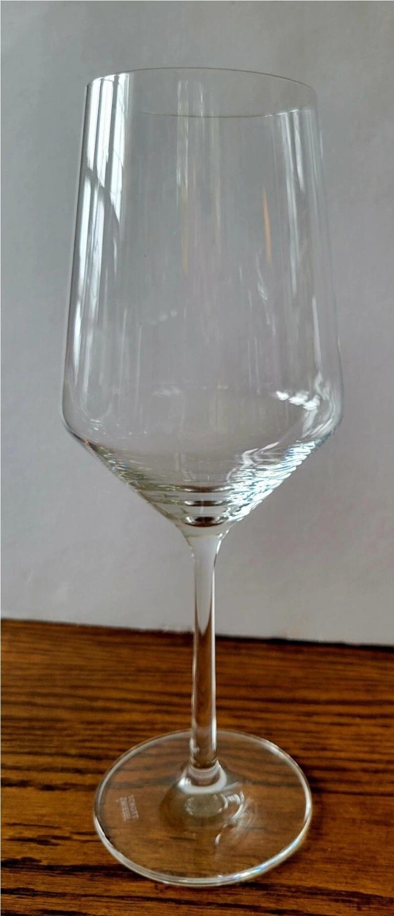 May include: A clear, stemmed wine glass with a wide, slightly tapered bowl and a thin stem. The glass sits on a round base. The glass is empty and reflects the light.