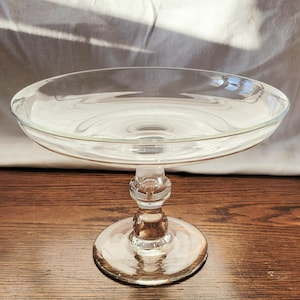 May include: A clear glass cake stand with a round base and a decorative stem. The stand has a wide, shallow bowl at the top. The stand is made of clear glass and sits on a wooden surface.