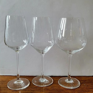 May include: Three clear wine glasses with different bowl shapes and long stems. The glasses are made of transparent glass and sit on a wooden surface. The glasses are designed for serving wine.