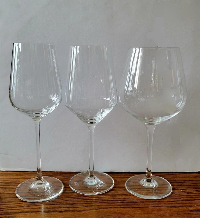 May include: Three clear wine glasses with different bowl shapes and long stems. The glasses are made of transparent glass and sit on a wooden surface. The glasses are designed for serving wine.