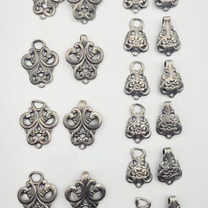 Könnte beinhalten: Eine Sammlung von silberfarbenen Metallschmuckkomponenten, die in zwei Spalten angeordnet sind. Die linke Spalte zeigt acht verzierte, symmetrische Schnörkelanhänger, während die rechte Spalte acht kleinere, blumenförmige Metallanhänger enthält.