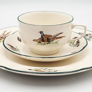 Puede incluir: Un juego de té de cerámica color crema compuesto por una taza, un plato y un plato grande. Cada pieza presenta un borde verde oscuro e ilustraciones detalladas de faisanes en colores naturales. La taza tiene un asa y forma redondeada.