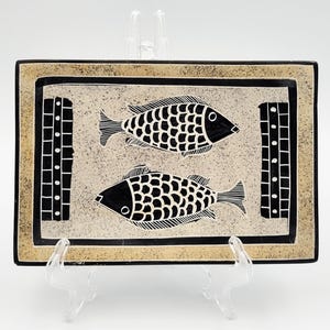 May include: A rectangular ceramic tray featuring a speckled beige background with a black border. Two black fish with white scale patterns are centered on the tray, flanked by two vertical black rectangular panels with white dot and grid details.
