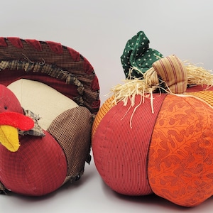 May include: A fabric turkey figurine with a red head, yellow beak, and brown checkered body, positioned next to a stuffed fabric pumpkin featuring orange, red, and yellow panels with a green stem and raffia accents for seasonal decor.