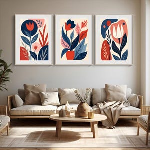 Può includere: Tre stampe artistiche floreali astratte incorniciate. Ogni stampa presenta fiori e foglie stilizzati in rosso, blu e rosa su uno sfondo color crema. Le stampe sono esposte sopra un divano in un soggiorno.