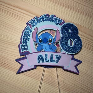 May include: A layered cake topper featuring the character Stitch from Lilo & Stitch. The topper has a blue glitter number 8, the name "ALLY", and the words "Happy Birthday". The design includes shades of blue, purple, and pink.