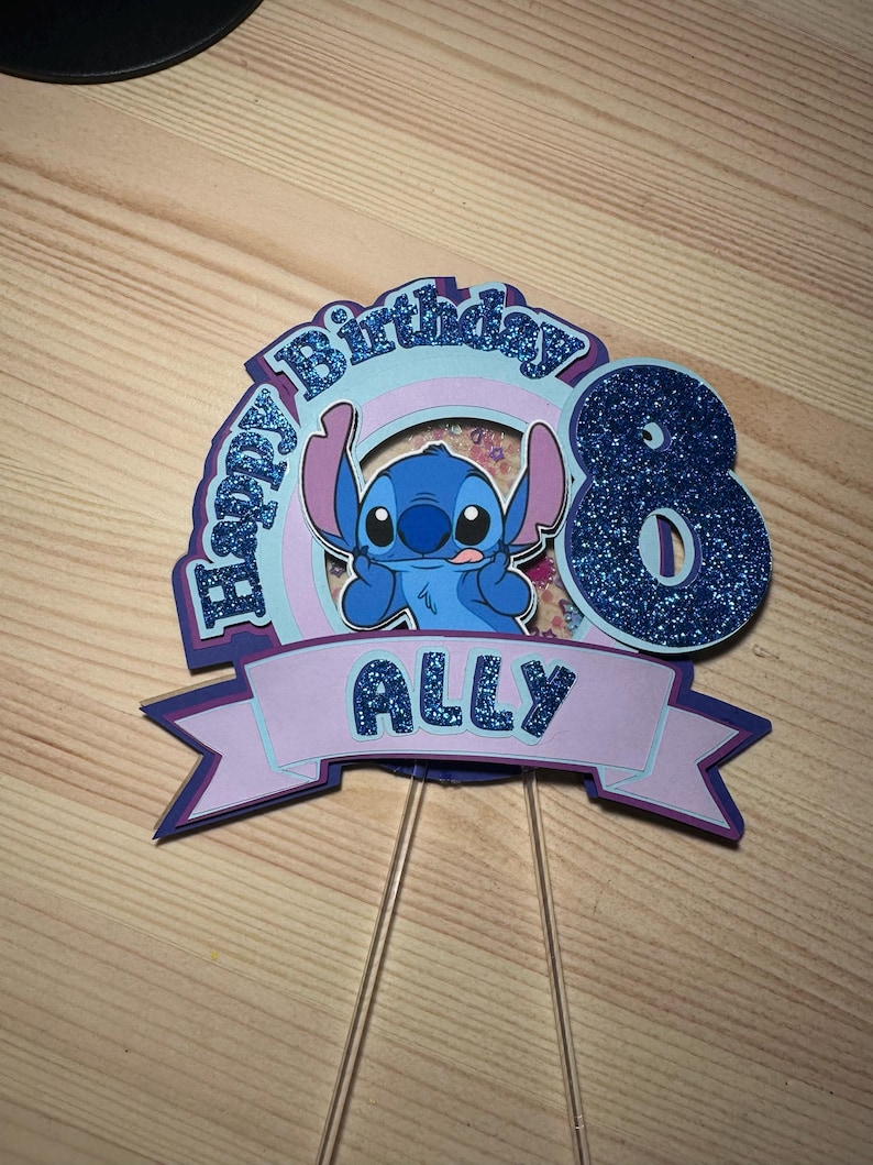 May include: A layered cake topper featuring the character Stitch from Lilo & Stitch. The topper has a blue glitter number 8, the name "ALLY", and the words "Happy Birthday". The design includes shades of blue, purple, and pink.