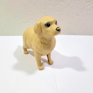 May include: A tan-colored plastic toy dog, resembling a Golden Retriever, stands on a white surface. The dog has blue eyes and a black nose. The toy is a solid color with a smooth texture, and is a children's toy.
