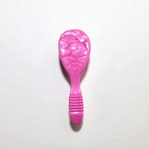 May include: A bright pink hairbrush with a floral design on the brush head and a ribbed handle. The brush is made of plastic and is set against a white background.
