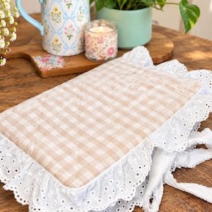 May include: A rectangular fabric cover featuring a beige and white gingham pattern, bordered by a wide white eyelet lace ruffle. The cover includes attached fabric ties at the corner, designed with a soft, quilted texture and intricate floral-patterned embroidery along the scalloped edges of the lace trim.