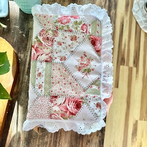 May include: A rectangular fabric cover featuring a patchwork design of pink roses, green stripes, and small floral patterns on a white background, finished with a white eyelet lace trim along the edges and a quilted texture throughout.
