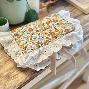 May include: A rectangular quilted fabric cover featuring a floral pattern with orange, yellow, and green flowers on a white background. The cover is edged with a wide, white eyelet lace ruffle and includes a light beige fabric ribbon tie.