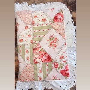 May include: A rectangular patchwork pillow featuring a mix of floral rose patterns and sage green stripes. The fabric is quilted with a white eyelet lace ruffle trim around the edges. The design includes small red bows and pink rose motifs.