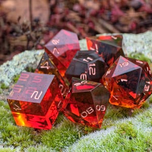 Può includere: Set di sette dadi poliedrici rossi traslucidi con numeri dorati, per giochi da tavolo. I dadi sono appoggiati su muschio verde.