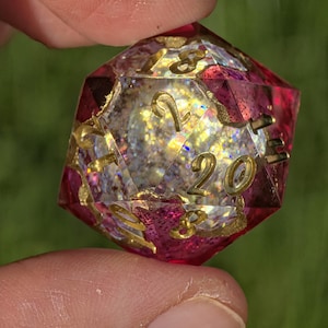 Può includere: Un dado D20 sfaccettato, rosa e oro. Il dado ha numeri dorati e un interno scintillante. Il dado è tenuto tra due dita su uno sfondo verde.