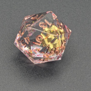 Könnte beinhalten: Ein klarer, vielseitiger D20-Würfel mit roségoldenen Zahlen und einem goldfarbenen Kern. Der Würfel ist mit schillerndem Glitzer gefüllt und vor einem dunkelgrauen Hintergrund platziert.