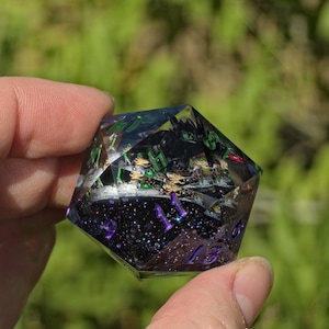 Könnte beinhalten: Ein zwanzigseitiger Würfel mit einer transparenten Harzhülle und einem dunklen, glitzernden Inneren. Der Würfel enthält eine kleine schwarze Drachenfigur und zeigt Zahlen in violetter Schrift, darunter die sichtbaren Zahlen 9, 11 und 13.