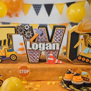 Puede incluir: Decoraciones de cumpleaños con temática de construcción con el nombre "Logan". Incluye recortes de números y letras amarillas y negras, camiones de juguete, conos de tráfico y cupcakes. Globos y una pancarta completan la fiesta.