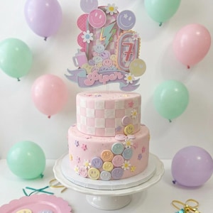 Puede incluir: Un pastel rosa de dos niveles decorado con un patrón de cuadros, pequeñas estrellas, flores y detalles de caras sonrientes. Un gran adorno para pastel muestra el nombre Piper, el número 7, un rayo y varios iconos de caras sonrientes pastel.