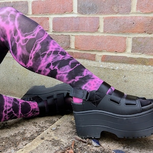 May include: Black platform sandals with multiple straps are paired with leggings featuring a vibrant pink and black lightning bolt pattern. The sandals have a thick sole, and the leggings create a bold, eye-catching look.