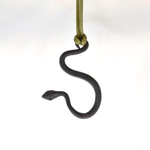 May include: A dark gray metal snake pendant hanging from a green cord. The snake is stylized with a curved body and a small, detailed head. The background is plain white.