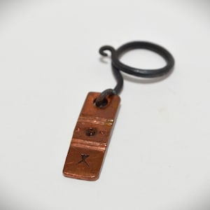 May include: A copper-colored pendant with a rectangular shape and a small, dark X symbol at the bottom. The pendant is connected to a black, circular ring via a curved wire. The pendant has a rustic, handmade appearance.