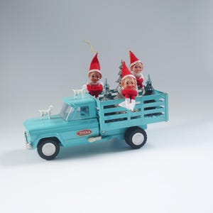 May include: A blue toy truck with a white Tonka logo on the side. The truck bed is filled with three small Christmas elves and silver tinsel trees.