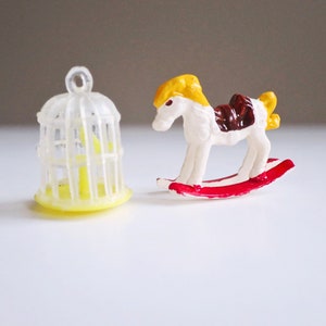 A three quarter inch yellow clear plastic bird cage gum1ball charm and 3/4 plastic rocking horse figurine for crafts or dollhouse decorations or toys.  The rocking horse is white with a yellow mane and red gliders.  More https://bit.ly/SO11vintage