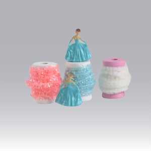 May include: Three spools of decorative trim, each with a different color and texture. The spools are pink, white, and blue. The blue spool has a small plastic figurine of a woman wearing a blue dress on top of it.