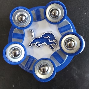 May include: A blue and silver fidget spinner with six silver ball bearings. The center features a blue and white logo of a lion. The spinner is on a dark surface.