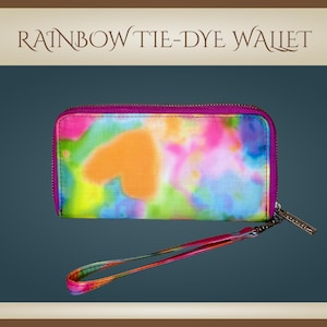 May include: Rainbow tie-dye wallet with a vibrant, colorful design. The wallet features a zipper closure, a wrist strap, and a bright pink trim. The tie-dye pattern includes shades of orange, pink, blue, and yellow.