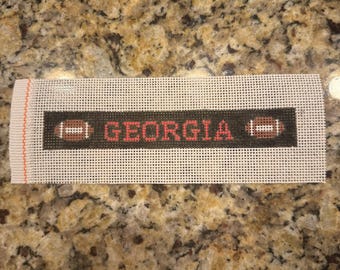 May include: A needlepoint canvas with the word "GEORGIA" in red letters. The word is flanked by two brown footballs with white laces. The background is black. The canvas is white with a grid pattern.