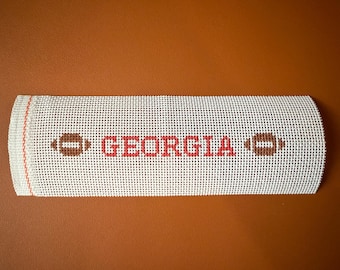 May include: A rectangular needlepoint canvas with the word "GEORGIA" in red block letters. Brown footballs flank the text. The canvas is white with a grid pattern and a red border on one end. The background is brown.