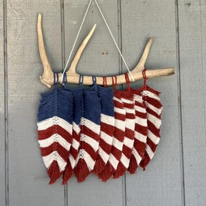 May include: A decorative wall hanging featuring a natural antler base with macrame feathers. The feathers are in a patriotic design, with alternating red and white stripes and blue sections, creating a unique rustic aesthetic.