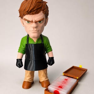 Op de afbeelding: Een gestileerd vinylfiguur van een persoon met kort bruin haar, gekleed in een groen shirt, een zwart schort en zwarte handschoenen. Naast het figuur staat een open bruine rechthoekige koffer met een rij doorzichtige glaasjes met rode stippen.