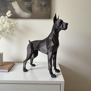 Puede incluir: Una escultura geométrica negra de un gran danés. La estatua del perro tiene un diseño facetado, con ángulos agudos y una estética moderna. La escultura está sobre una superficie blanca, con un fondo neutro.