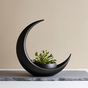 May include: A black, crescent-shaped planter with a small, round pot nestled inside, holding green plants. The planter sits on a gray, rectangular surface. The design is modern and minimalist.