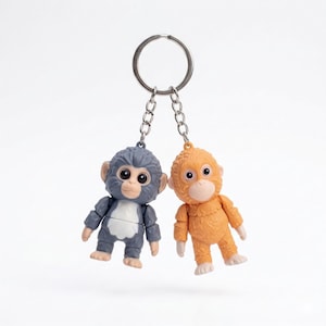 Peut inclure: Deux porte-clés singes. L'un est gris et blanc, l'autre orange. Les deux ont de grands yeux et sont attachés à un porte-clés argenté avec une chaîne. Les porte-clés sont de petites figurines détaillées.