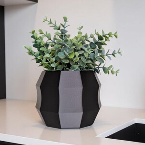 Puede incluir: Un jarrón decorativo geométrico negro y gris que contiene una planta verde con hojas. El jarrón tiene un diseño facetado y se encuentra sobre una encimera blanca. La planta tiene hojas verdes pequeñas y redondas.
