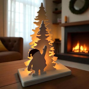 May include: A white, illuminated Christmas tree and Santa Claus silhouette decoration. The tree is layered with a cutout design, and the Santa figure is waving. The base is a white rectangle, and the warm light shines through the tree.
