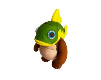 May include: A quirky figurine depicting a fish-like character. It features a green head with yellow fins, blue eyes, and brown limbs. The body is a light tan colour, and the design has a cartoonish aesthetic.