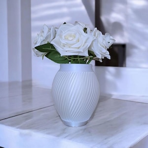 May include: A white vase with a spiral ribbed design holds a bouquet of white roses with green leaves. The vase sits on a white marble surface, creating a clean and elegant aesthetic. The arrangement is a classic home decor item.
