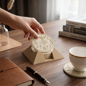 May include: A set of circular beige coasters featuring engraved Egyptian symbols including the Eye of Horus, a scarab, and an ankh. The coasters are stored in a pyramid-shaped holder, with one coaster being lifted to reveal the detailed carvings.