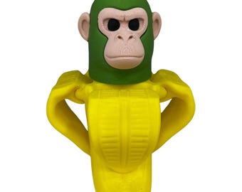 May include: A novelty banana-shaped object with a green monkey head. The monkey's face is a light tan colour, with black eyes and a detailed facial structure. The banana is yellow with a textured surface and a square base.