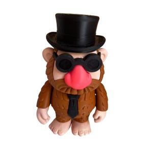 May include: A whimsical figurine of a troll-like character, sporting a black top hat and round sunglasses. The figure has a brown body, a pink nose, and a black tie. The design is playful and unique.
