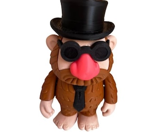 May include: A novelty figurine of a troll-like character, wearing a black top hat and round sunglasses. The figure has a brown body, a pink nose, and a black tie. The design is quirky and fun.