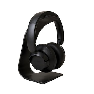 May include: Black over-ear headphones resting on a matching black headphone stand. The headphones have large earcups and a padded headband. The stand has a curved design with a base for stability. The headphones appear to be wireless.